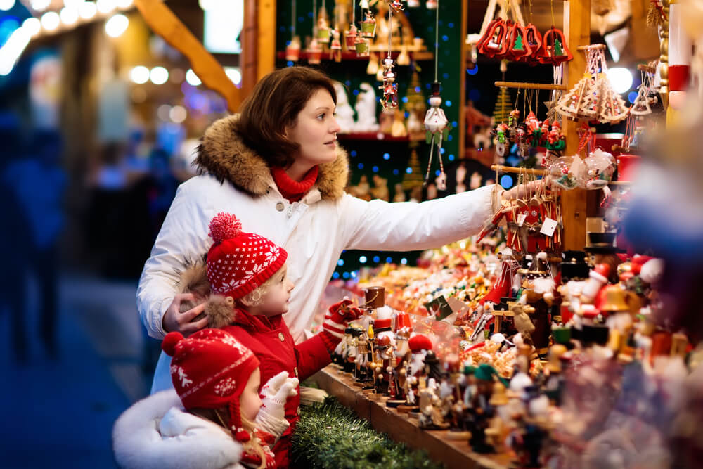 Mother and child in warm hat watching handmade glass Christmas tree ornaments at traditional German Xmas street market. Family with kids shopping for Christmas presents on winter fair on snowy day.