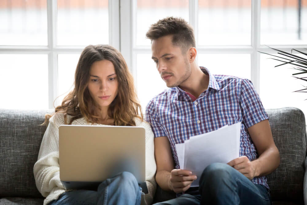 worried couple paying bills online on laptop with documents sitting together on sofa at home